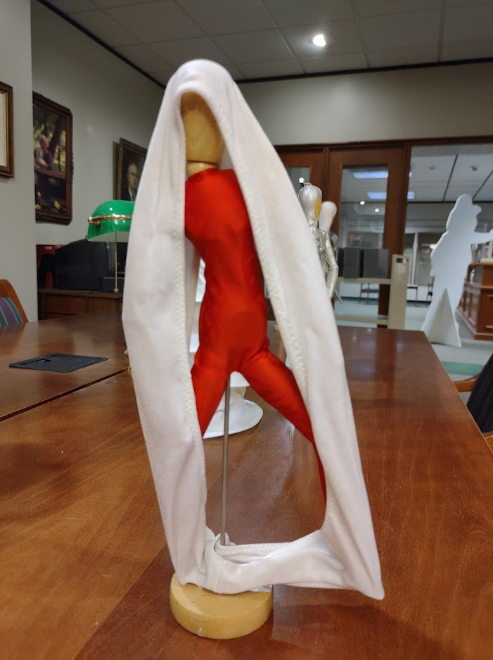 A small wooden 3-d figure wears a red body stocking and is encircled by a white cloth. 