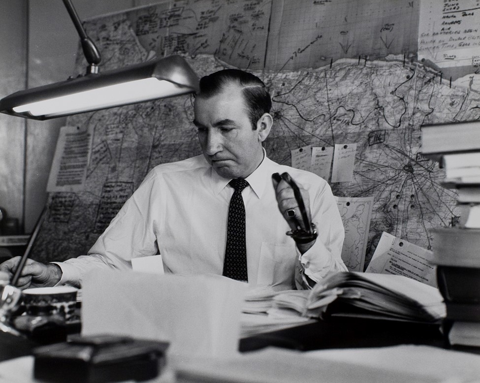 Cornelius Ryan sitting at his desk with a large map behind him.