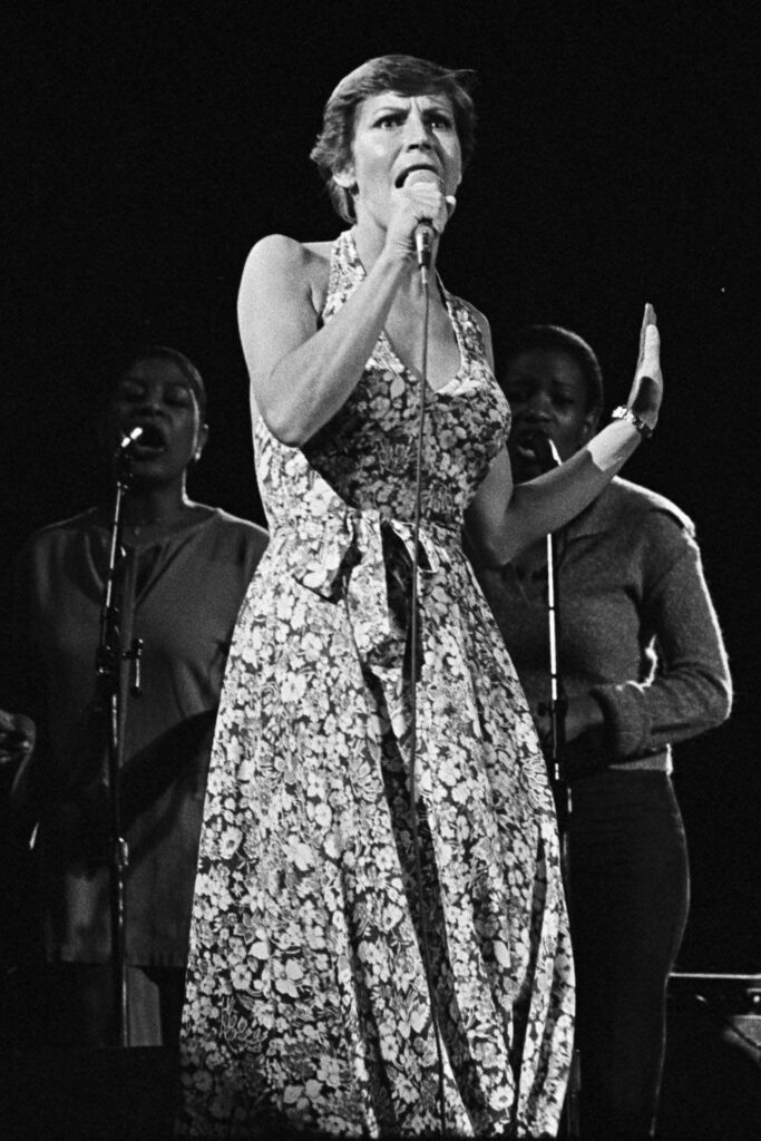 Helen Reddy in a floral dress singing into a microphone on stage in 1978