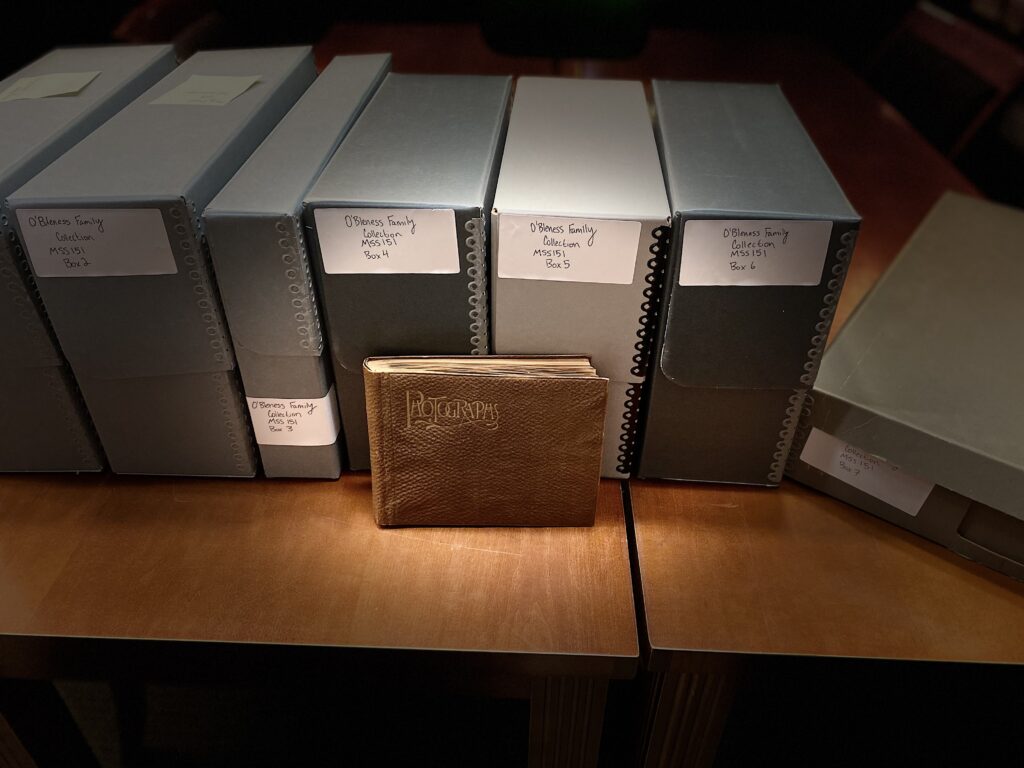 Archival boxes labeled "O'Bleness Family Collection" sit on a table with a photograph album leaning on them.