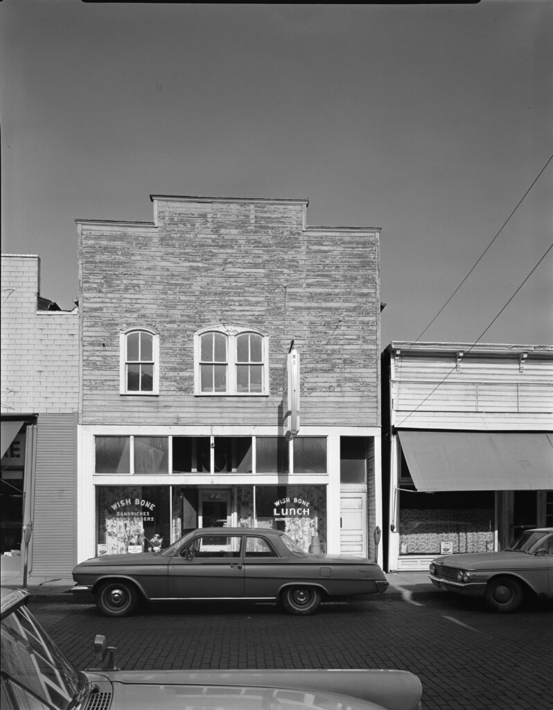 Wish Bone restaurant and neghboring storefronts on street with a vehicle parked in front, circa 1968-1969