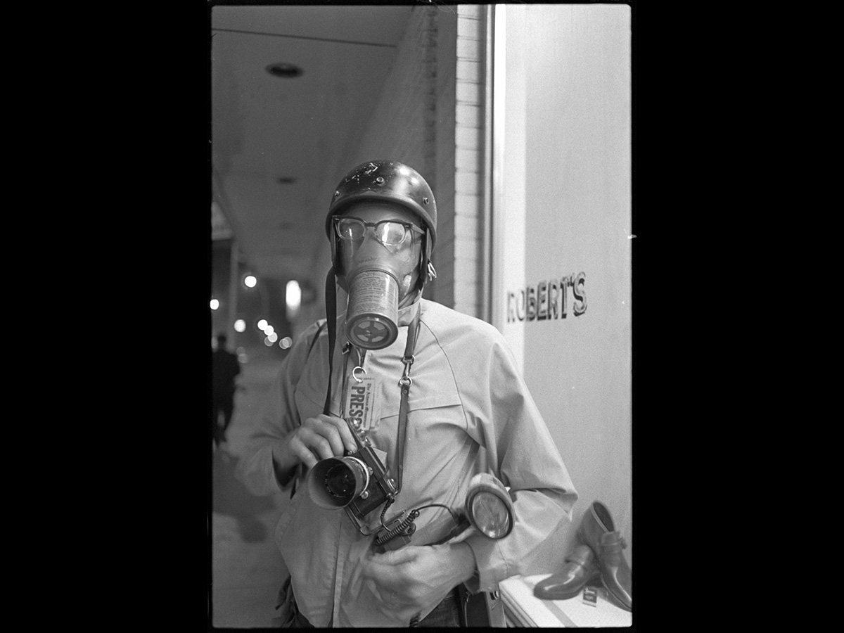 An Athens Messenger press photographer wearing a gas mask and riot helmet stands with camera in hand on a sidewalk at night
