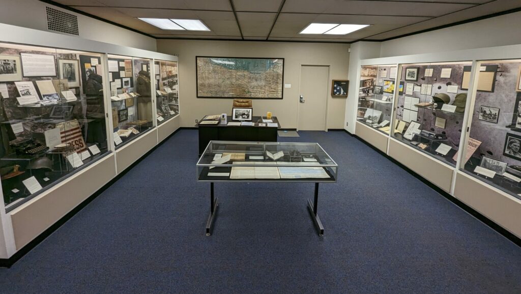 wide angle view of new exhibition with large display cases on the right and left walls, a flat display case in the center, and a desk and large map at the back of the room.