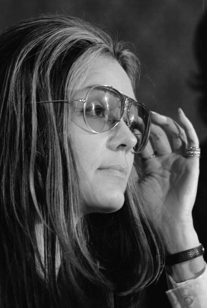 Black and white closeup snapshot of Gloria Steinem in profile wearing lightly tinted sunglasses and reaching hand toward face