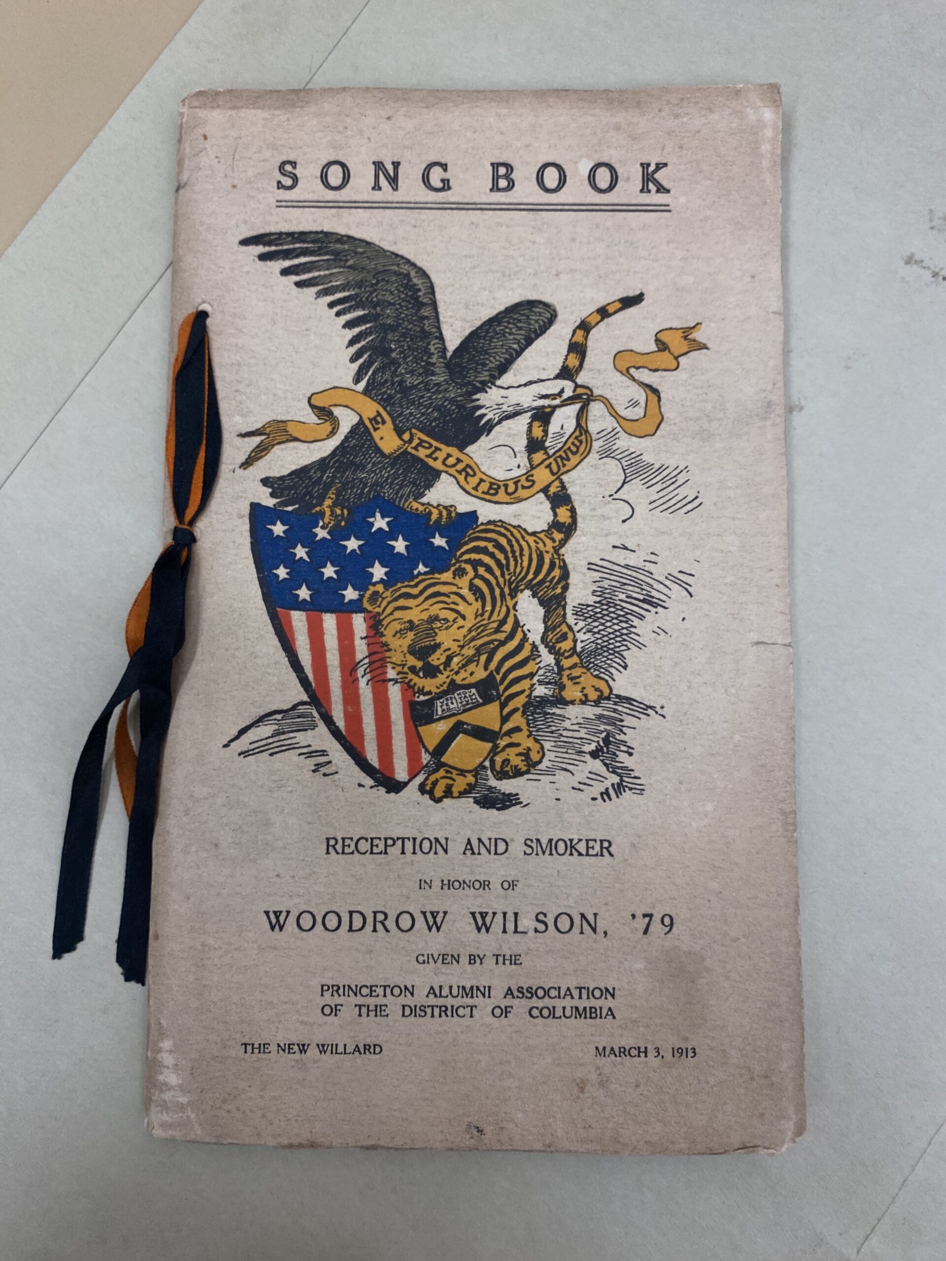 A song book held together with a ribbon, has a bird, lion, and US flag shield color illustration on the cover.