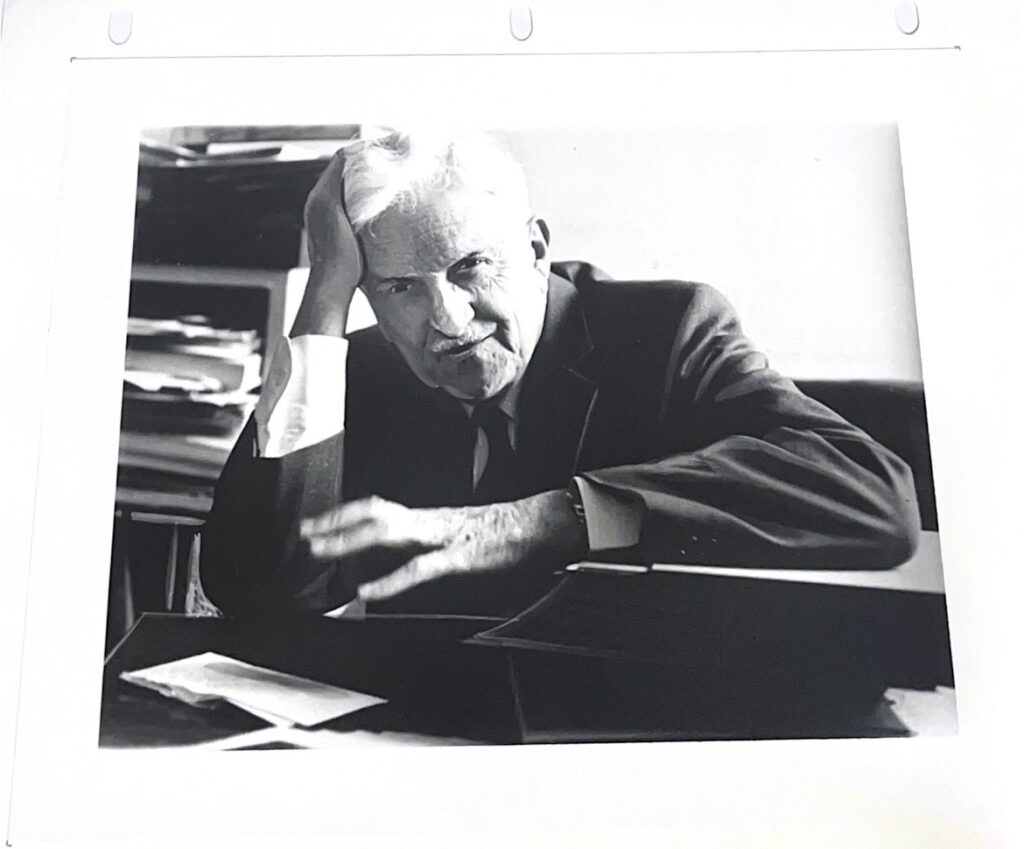 Black and white portrait of George Starr Lasher sitting at a desk with one hand touching his head and the other blurred as if moving on his desk.