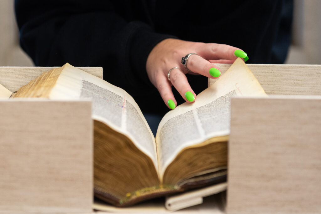 A student with green-painted fingernails turns a page in a very thick, old bible that sits in a cradle.