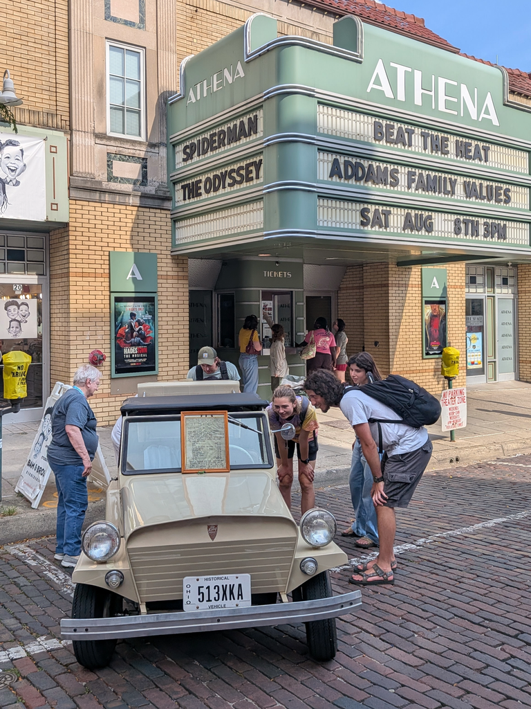 Five people stand around a parked tan King Midget car looking at it. In the background is Athena Cinema.