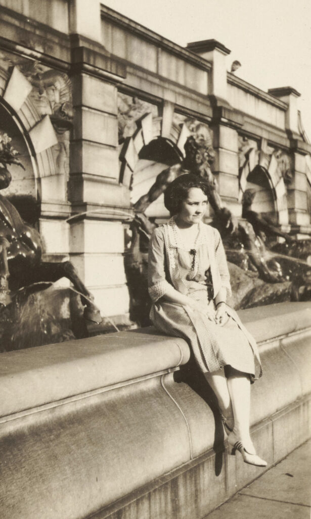 Black and white photo of Marie Deffenbaugh seated on the edge of an ornate fountain and wearing a checkered dress with bob-styled hair.
