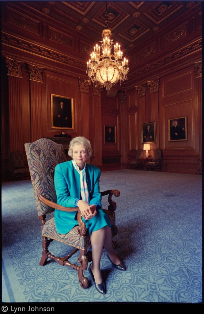 Seated portrait of Sandra Day O’Conner wearing a teal skirt suit in a sparsely furnished, ornate ballroom with a chandelier overhead and architectural detailing on the walls and ceiling.