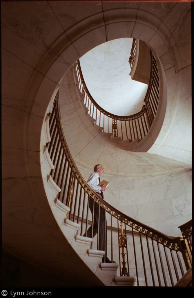 Man with a book in hand descends a grand spiral staircase made of marble with decorative gold-detailed railing.