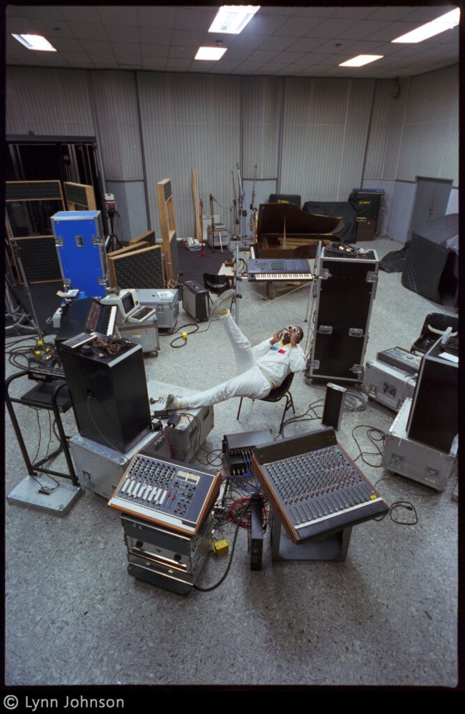 Overhead view of musician Stevie Wonder surrounded by audio equipment in a music studio, leaning back in a chair with one leg in the air while playing a harmonica.