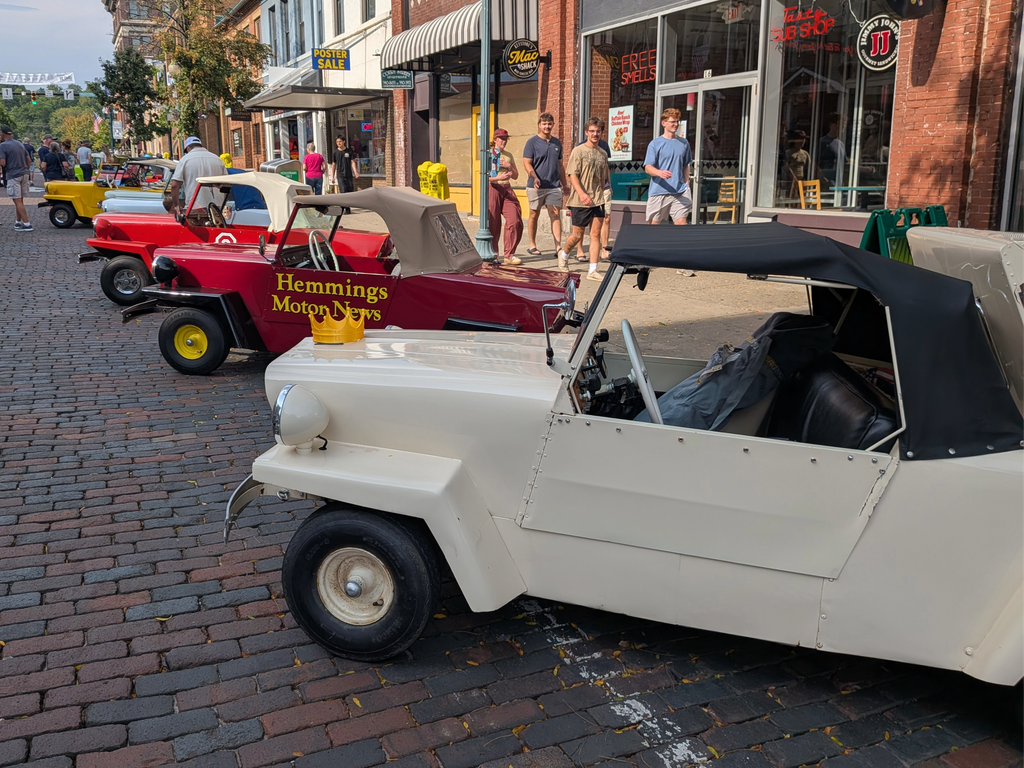 A white King Midget car parked on a brick street with a gold crown on its hood, next to a red King Midget car with "Hemmings Motor News" written on it in yellow, and people walking on a sidewalk in the background.