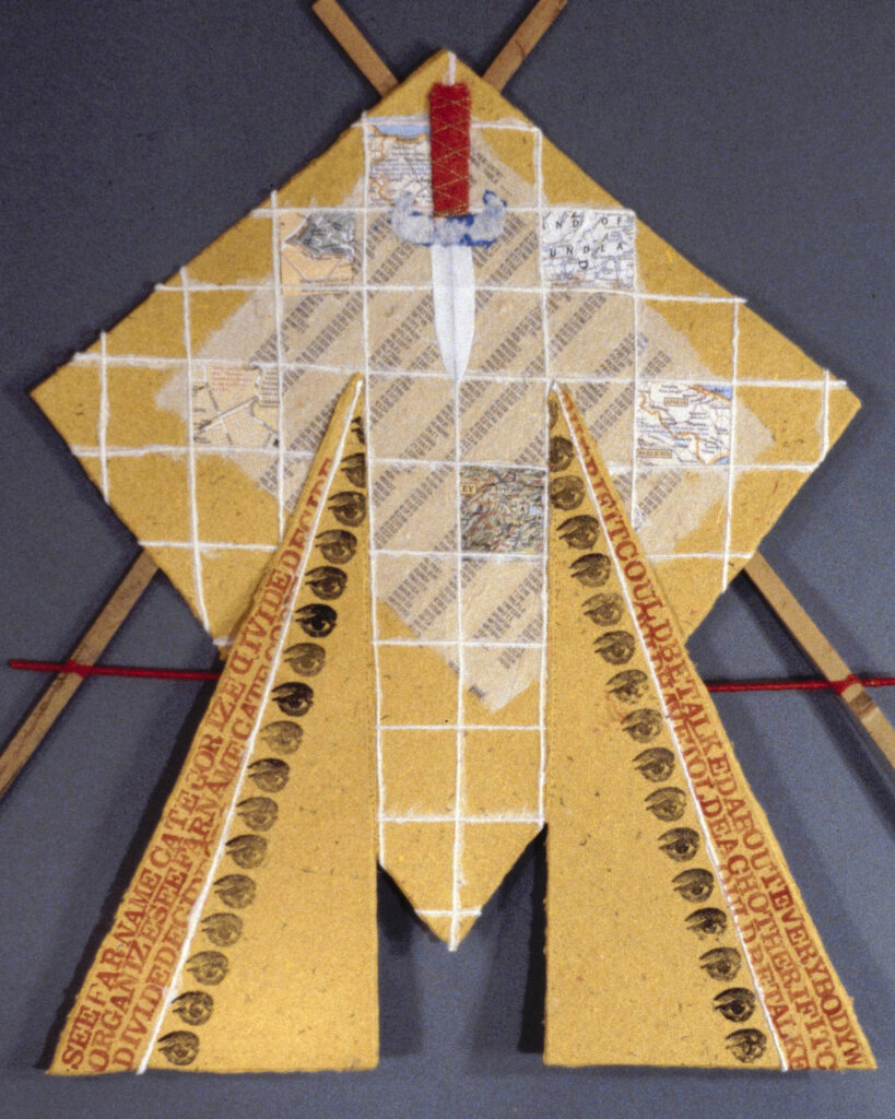 Art work incorporating yellow handmade paper, stamped text and eyes, collaged maps, and a cutout of a dagger, assembled with reeds and red string to form a shield.