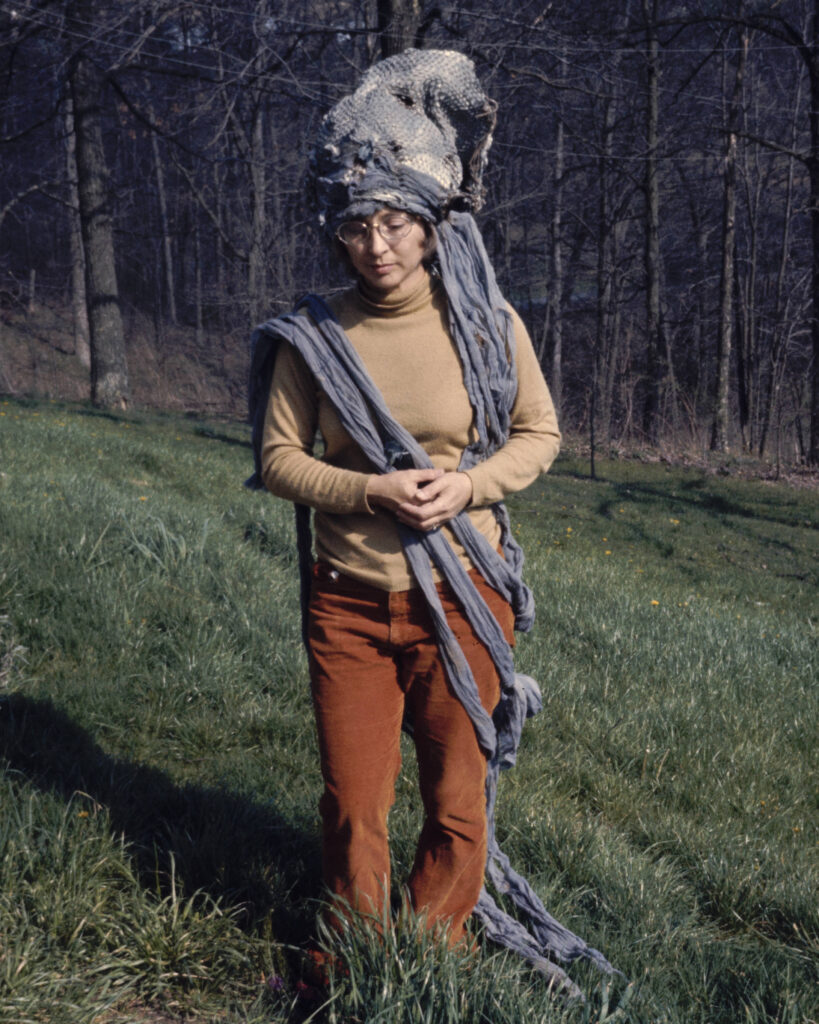 Outdoor portrait of Sara Gilfert with head bowed to model a slouched and netted gray fabric headpiece with extensions draped over her shoulder and lying in the grass.