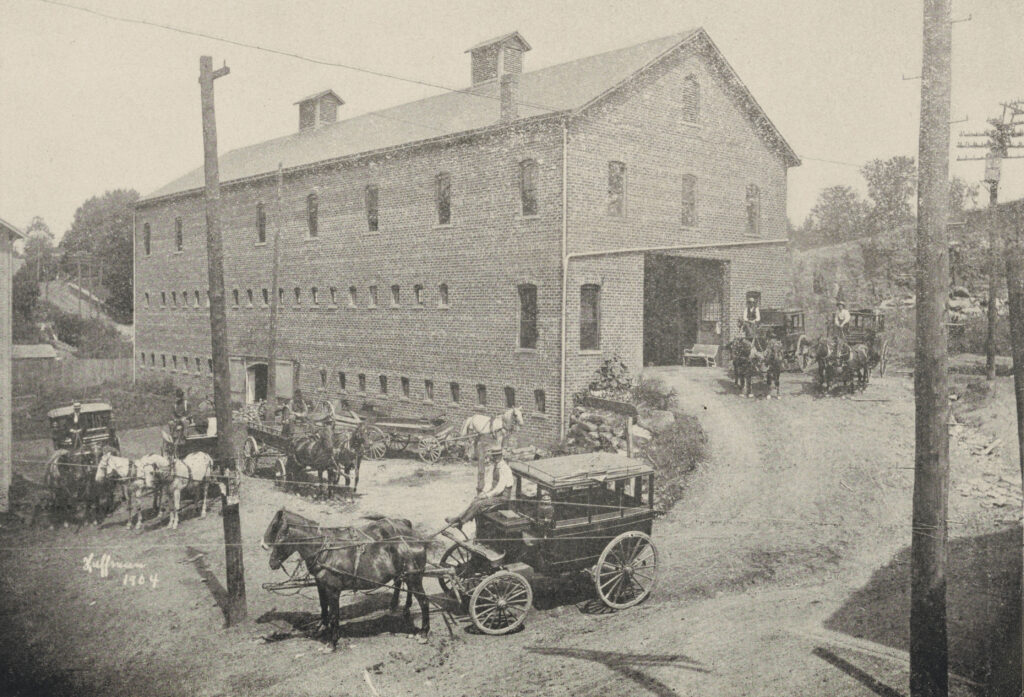 Black-and-white halftone printed photo of large brick livery structure with horse-drawn carriages outside.