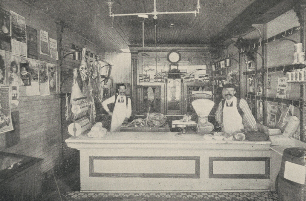 Black-and-white halftone printed photo of two men wearing aprons behind a counter with meats hanging along the wall.