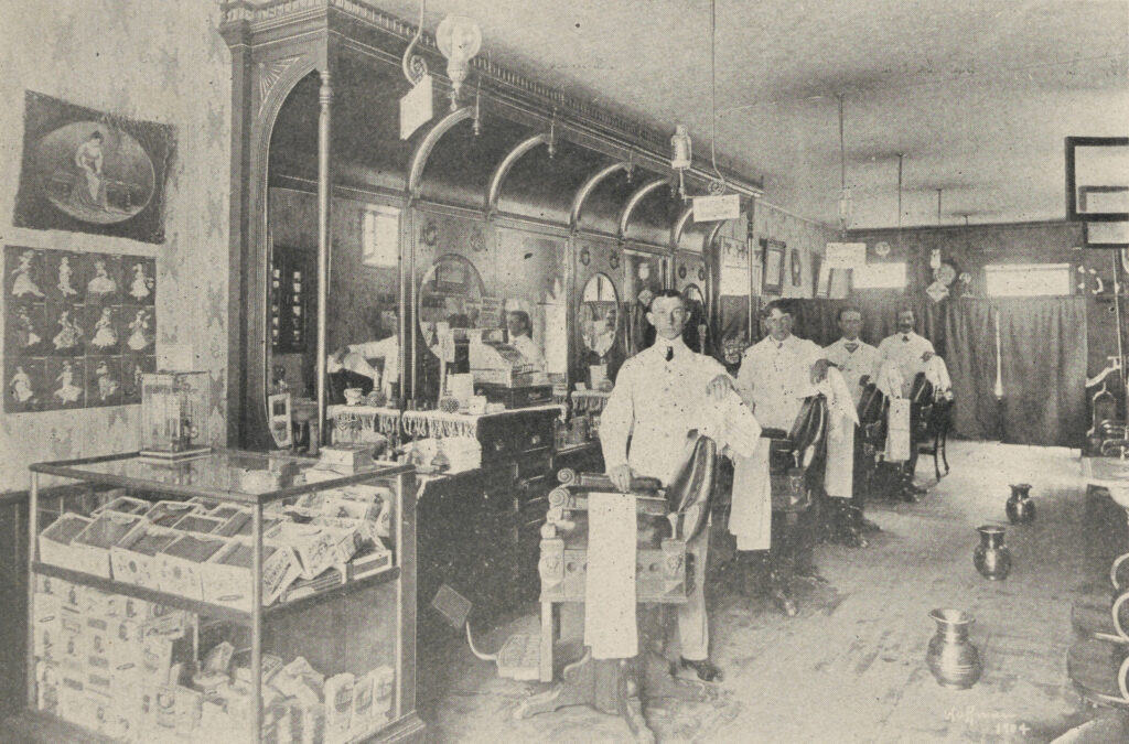 Black-and-white halftone printed photo of shop interior with four barbers standing beside chairs along a wall of vanities and mirrors.