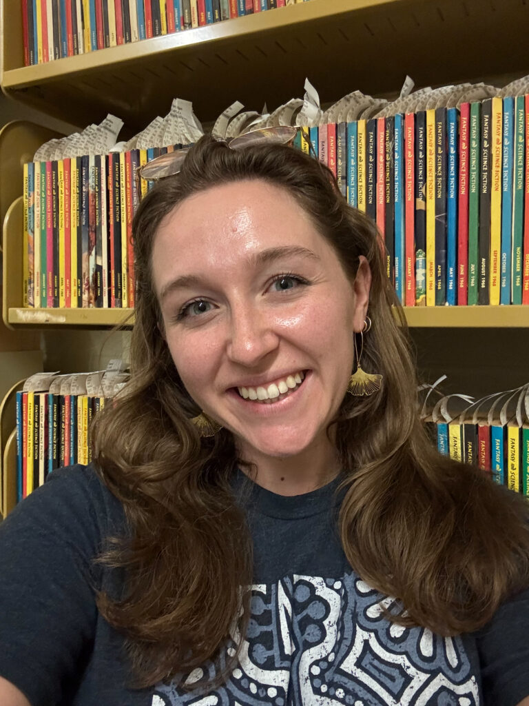 GA Mary Moore stands in front of multi-colored paperback magazines on a shelf with call number flags sticking out of the top. She has glasses tucked in her hear, is wearing a navy and white shirt, and is wearing gold ginkgo leaf earrings.