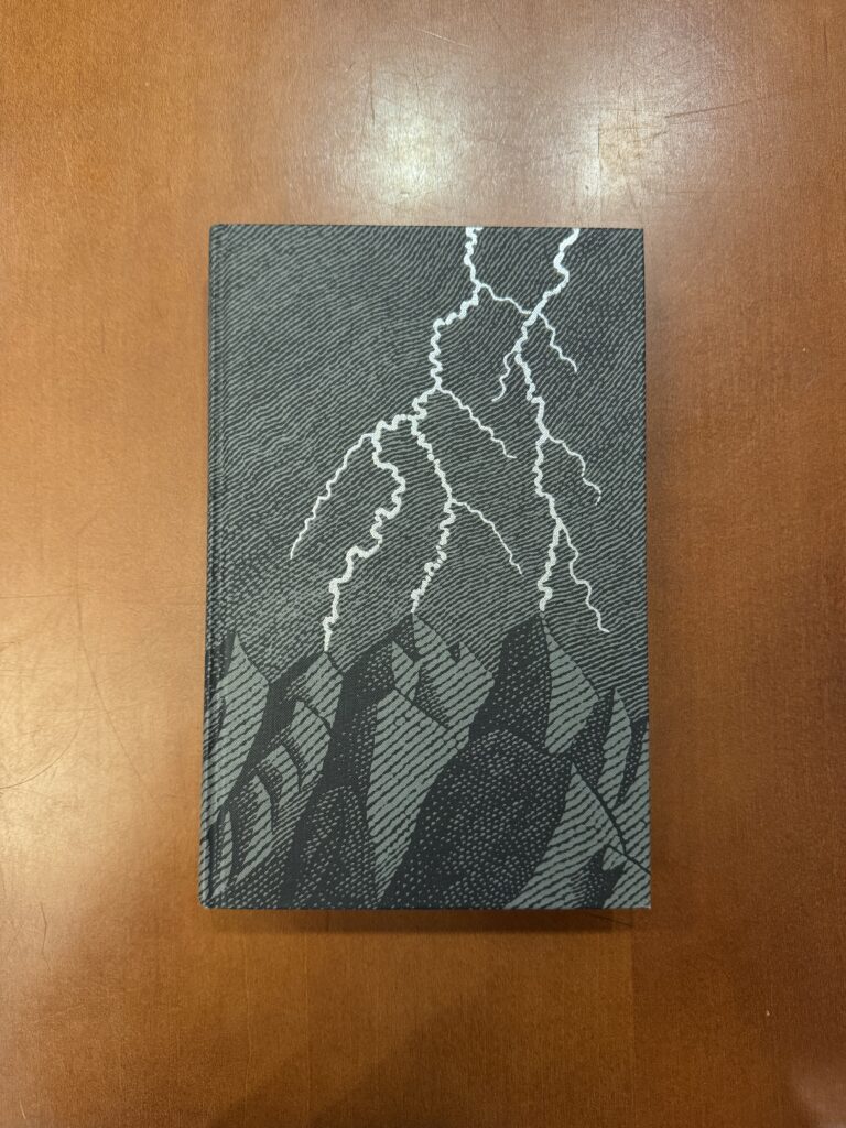 A dark grey book lies on a brown table. The cover features a silver embossed illustration of lightning over black, woodcut style drawings of mountains.  
