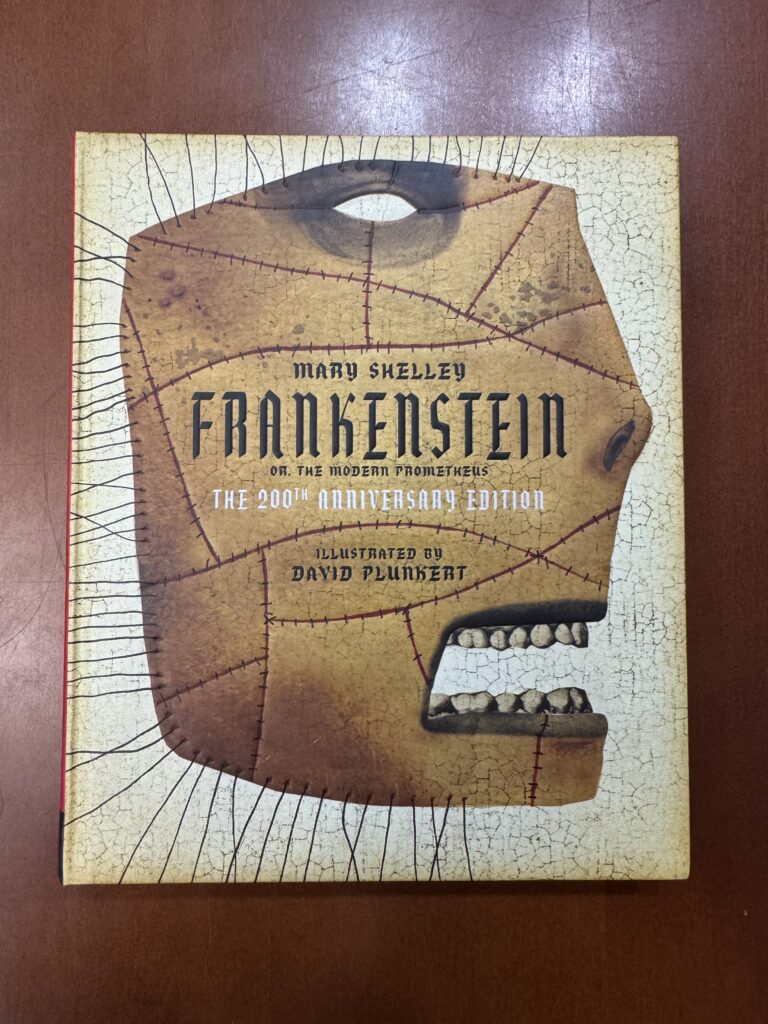 A white book lies on a brown table. Text on the cover reads: “Mary Shelley. Frankenstein or, the Modern Prometheus. The 200th Anniversary Edition. Illustrated by David Plunkert. The book cover features an illustration of a face with patchworked skin. 