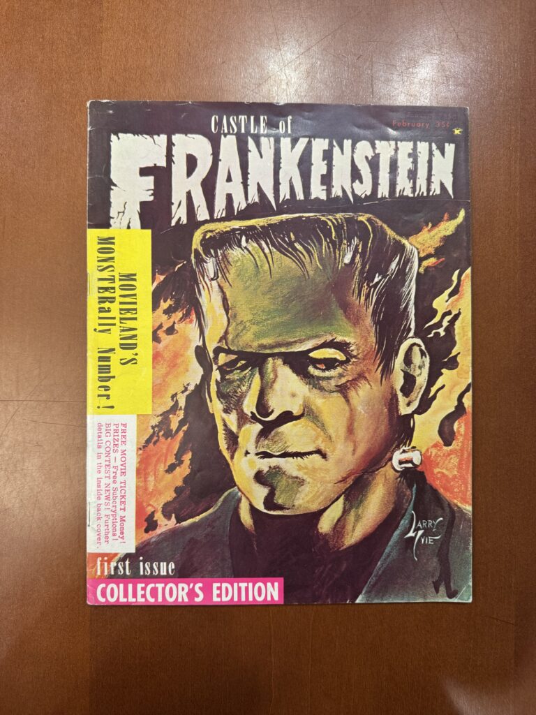 An illustrated magazine lies on a brown table and features a large illustration of a flat headed, green skinned Frankenstein’s monster. Text reads: “Castle of Frankenstein. February 35c. First Issue Collector’s Edition. Sideways advertisements along the spine of the magazine feature text that reads: “Movielands Monsterally Number!” and “Free Movie Ticket Money! Prizes — Free Subscryptions ! Big Contest News! Further details in the inside back cover.  