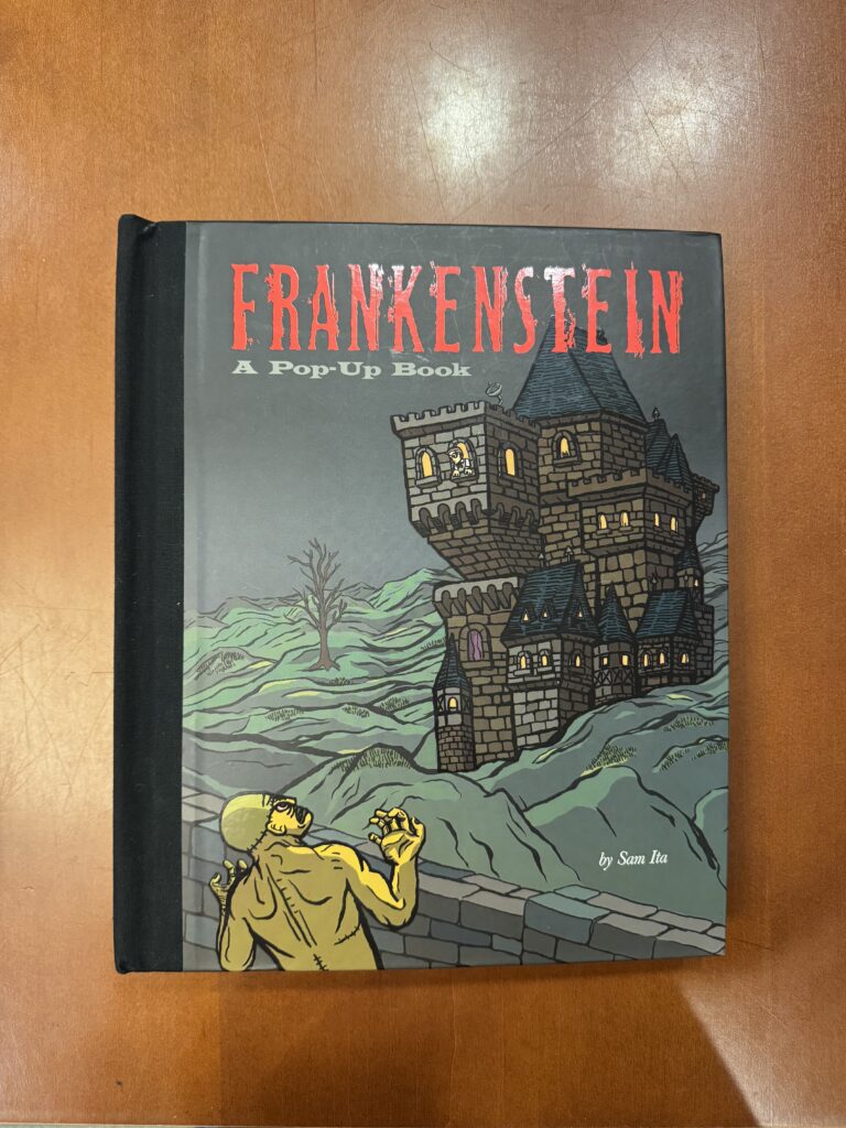 A grey book with a black spine lies on a brown table. It features an illustration of a stone castle on a hilly landscape with a green figure in the foreground. Text in red reads: “Frankenstein.” Additional text on the cover reads: “A pop-Up Book” and “by Sam Ita”  