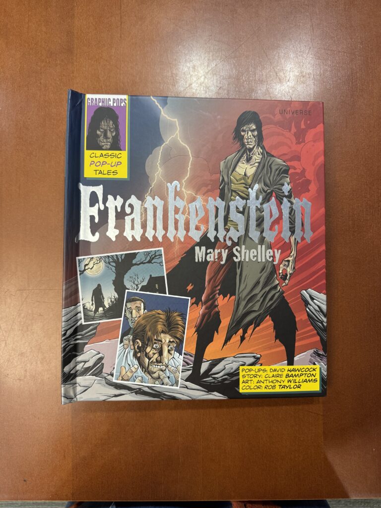 A highly illustrated book lies on a brown table. Text across the cover reads: “Graphic Pops. Classic pop-up tales. Frankenstein. Mary Shelley. Pop-ups: David Hawcock. Story: Claire Bampton. Art: Anthony Williams. Color: Rob Taylor. Illustrations include several images of a menacing figure.  