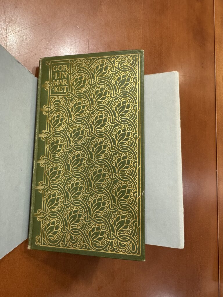 A green book lies in a grey cradle on a brown table. The cover is embossed with an elaborate gold decorative design and features the text: “Goblin Market.” 