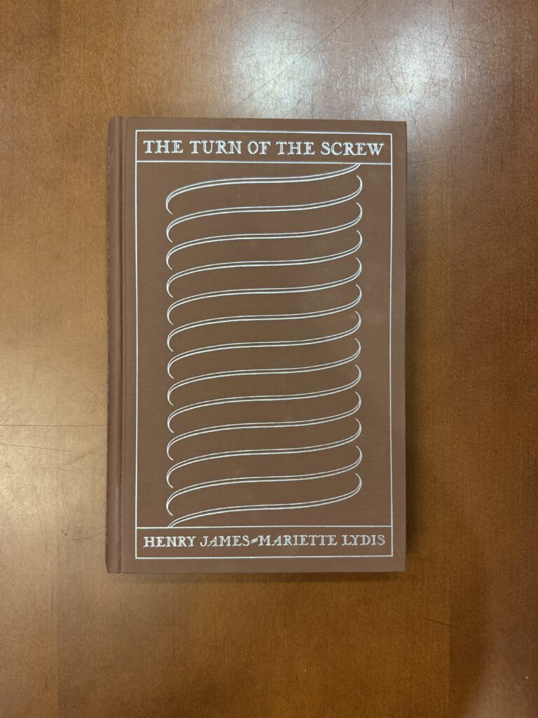 A grown book lies on a brown table. A silver embossed design takes up most of the cover. Text reads: “The Turn of the Screw. Henry James—Mariette Lydis.” 