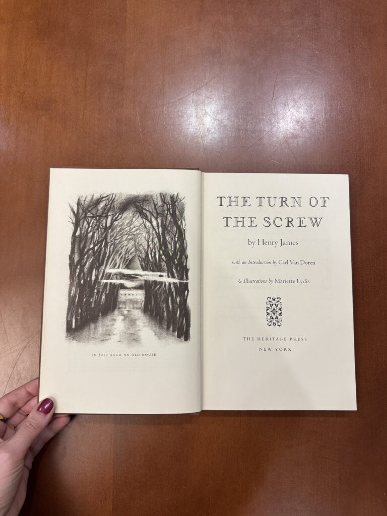 A book lies on open on a brown table, with a white hand holding the left book corner. The left page features a black and white illustration of a house with large looming trees. The right page features the text: “The Turn of the Screw. By Henry James. With an introduction by Carl Van Doren. & Illustrations by Mariette Lydis. The Heritage Press. New York.”  
