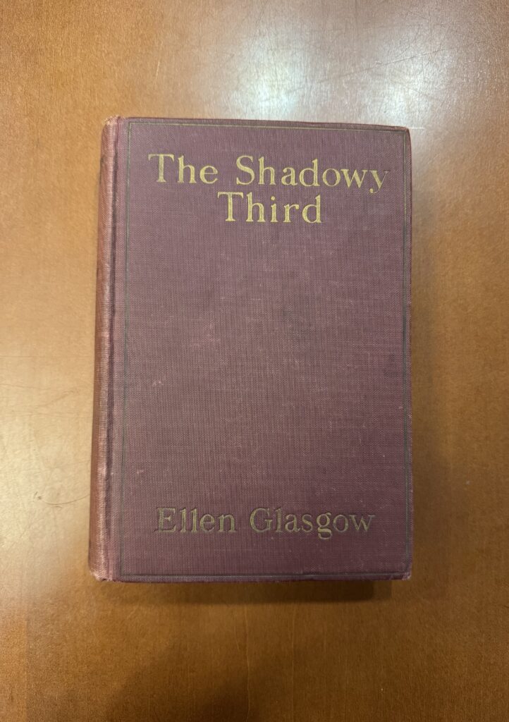 A maroon book lies on a brown table. Gold embossed text reads: “The Shadowy Third” and then “Ellen Glasgow.” 