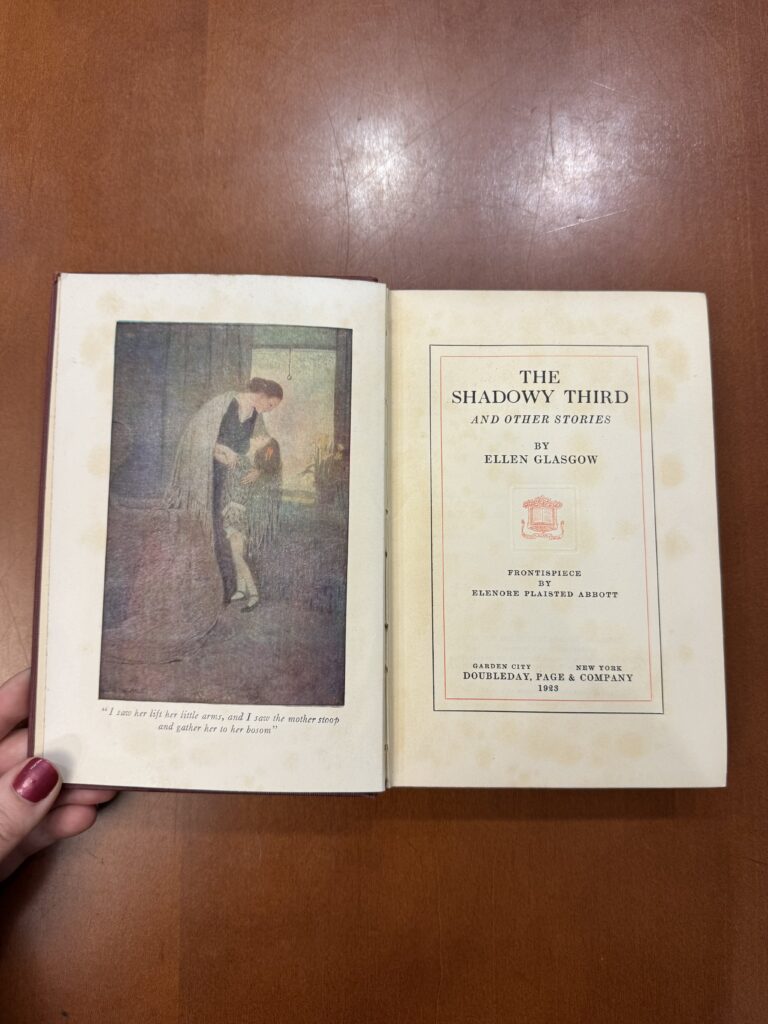 A book is held open on a brown table. On the left page there is a frontispiece illustration of a woman holding a young girl. On the right page text reads: “The Shadowy Third and Other Stories by Ellen Glasgow.  Frontispiece by Elenore Plaisted Abbott. Garden City. New York. Doubleday, Page & Company. 1923.  