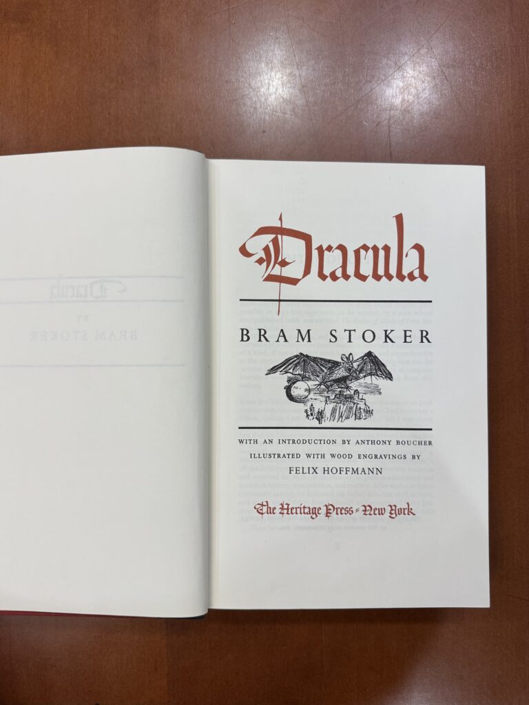 A book lies open on a brown table. The left page is blank, and the right page features an illustration of a bat in flight. Text reads: “Dracula. Bram Stoker. With an Introduction by Anthony Boucher. Illustrated with Wood Engravings by Felix Hoffmann. The Heritage Press New York.  