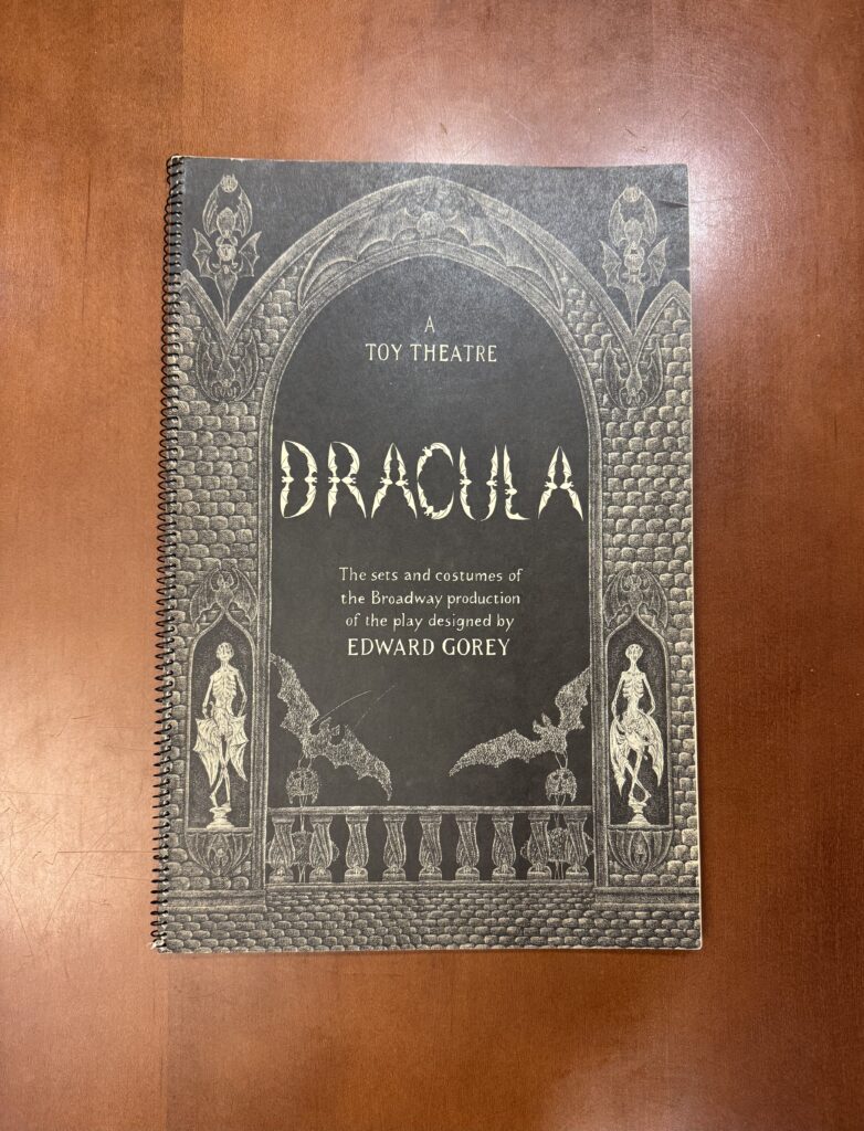 A large spiral bound book lies on a brown table. It is features highly detailed illustrations of a gothic style window. Text reads: “A Toy Theater. Dracula. The sets and costumes of the Broadway production of the play designed by Edward Gorey.”  