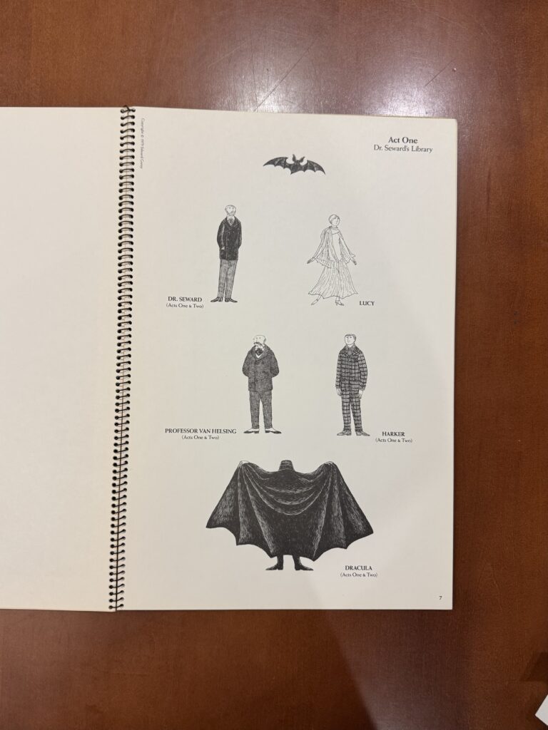 A spiral bound book lies open on a brown table. Features illustrations of five figures and one black bat.  