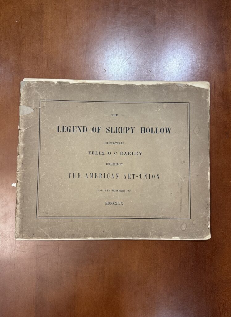 A large rectangular book lies on a brown table. The text on the cover reads: The Legend of Sleepy Hollow illustrated by Felix O C Darley Published by The American Art-Union for the Members of MDCCCXLIX.  