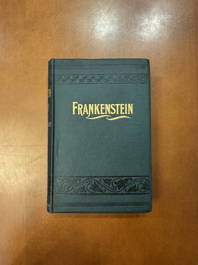  A green covered book lies on a brown table with “Frankenstein” embossed in gold w/ decorative florals and designs embossed in black ruling above and below the title across the cover.  