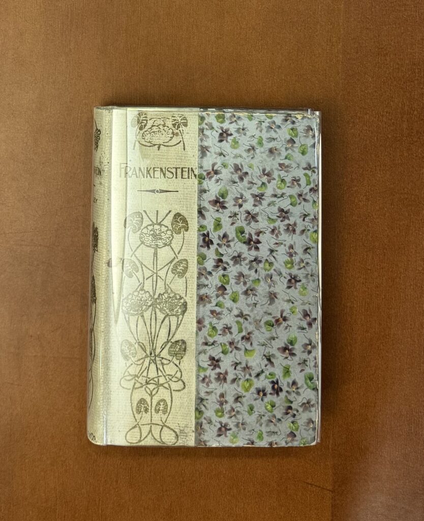  A book sits on a brown table; its cover is grey with purple flowers and green leaves. The spine is wrapped in cream paper and features the text “Frankenstein” printed in a serif font followed by an embellished floral design.  