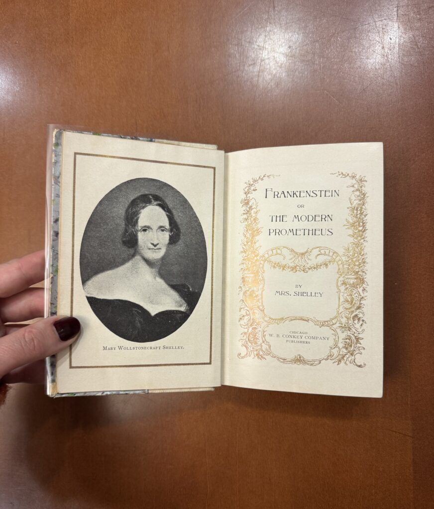 A white person’s hand with maroon nail polish holds open a book; on the left side is the portrait of a white woman in a black dress with bare shoulders, captioned with the text “Mary Wollstonecraft Shelley” in all caps, in a serif font. The right side features the text: “Frankenstein or The Modern Prometheus” in an all caps serif font: “by Mrs. Shelley” in a smaller size, same serif font, and “Chicago W. B. Conkey Company Publishers” in the same serif font, even smaller. The text on the page is surrounded by a gold embossed floral decoration. 
