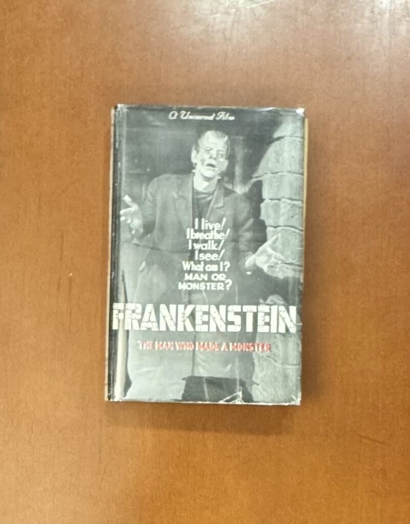 A book sits on a wooden table; the cover features a black and white image of a man dressed as Frankenstein’s monster. At the top of the cover script text reads: “A Universal Film.” Text on top of the image reads: “I live/I breath/I walk/I see/What am I? Man or Monster” in a sans serif font. Below that all large caps text, stylized with bolts, reads: “Frankenstein.” Below that in all caps, red text reads: “the man who made a monster.” 