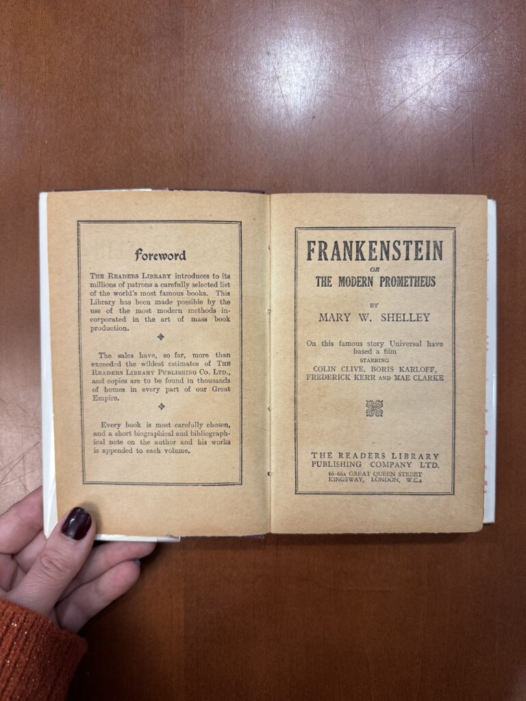 A white hand with maroon nail polish holds open a book; a small portion of an orange sweater covers the hand. The left page reads: “Foreword. The Readers Library introduces its millions of patrons a carefully selected list of the world’s most famous books. This Library has been made possible by the use of the most modern methods incorporated in the art of mass book production. The sales have, so far, more than exceeded the wildest estimates of The Reader's Library Publishing Co. Ltd. And copies are to be found in thousands of homes in every part of our Great Empire. Every book is most carefully chosen, and a short biographical and bibliographical note on the author and his words is appended to each volume. The right page features the text: “Frankenstein or The Modern Prometheus by Mary W. Shelley. On this famous story Universal have based a film starring Colin Clive, Boris Karloff, Frederick Kerr and Mae Clarke. The Readers Library Publishing Company LTD. 66-66a Great Queen Street Kingsway, London, W.C.2.  