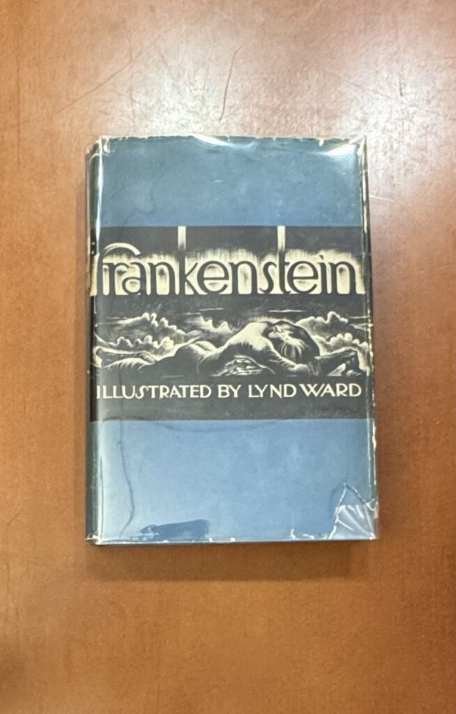 A book lies on a brown table: the cover is blue and wrapped in shiny plastic wrap. Lowercase text reads “frankenstein” above a black and white illustration of a figure crawling. Below the illustration all caps white text reads: “Illustrated by Lynd Ward”  