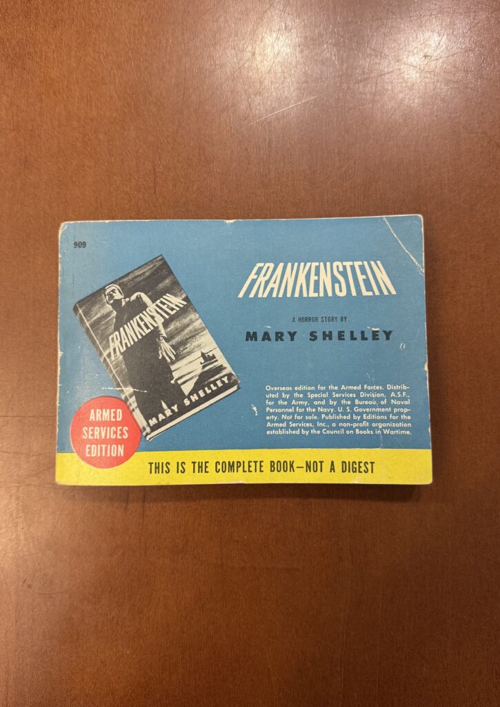 A small blue book lies on a brown table. The cover features the text “909. Frankenstein. A horror story by Mary Shelley. Overseas edition for the Armed Forces. Distributed by the Special Services Division, A.S.F. for the Army, and by the Bureau of Naval Personnel for the Navy, U.S. Government Property. Note for Sale. Editions for the Armed Services Inc., a non-profit organization established by the Council of Books in Wartime.” In a red circle white text reads: “Armed Services Edition.” A yellow stripe at the bottom of the cover reads “This is a complete book —not a digest. The cover also features an image of the book Frankenstein with a black and white cover and an image of the creature. 